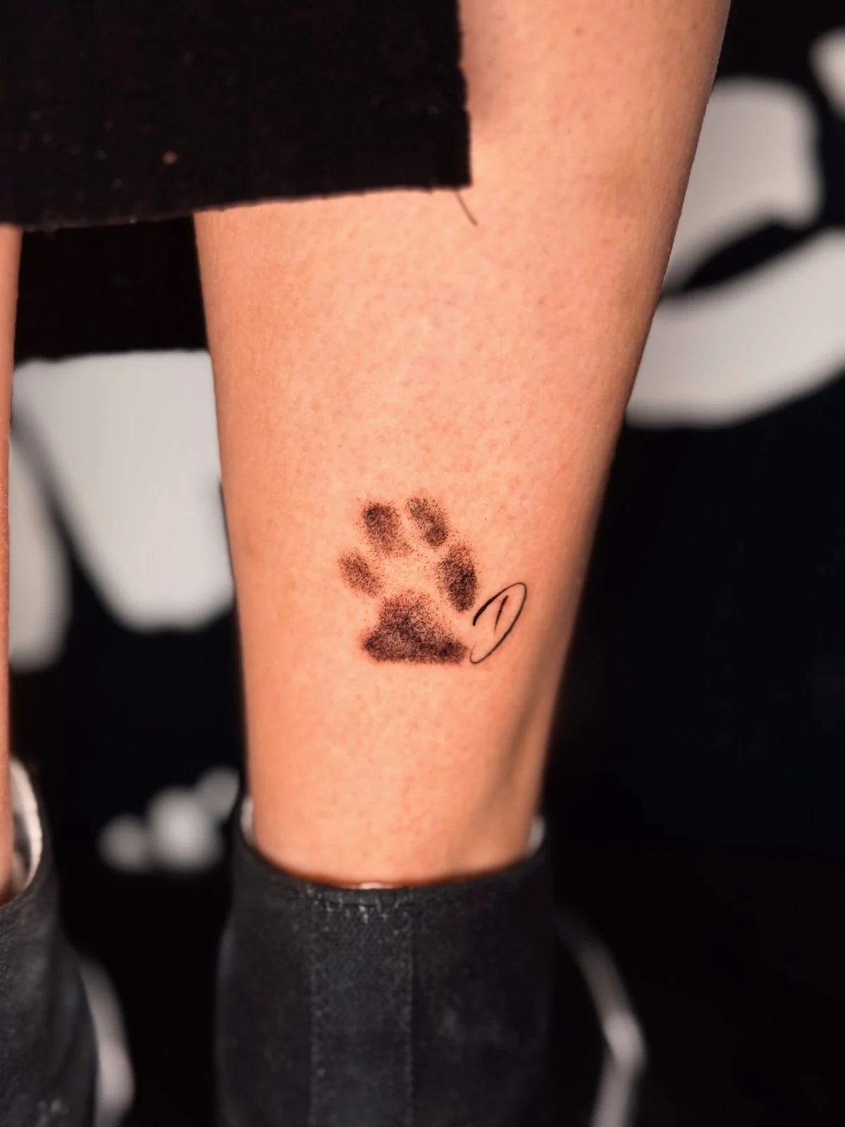 Kleines Pfotenabdruck-Tattoo in Dotwork-Technik mit einem geschwungenen Buchstaben am Bein – Arbeit von Merci, Skin City Tattoo.
