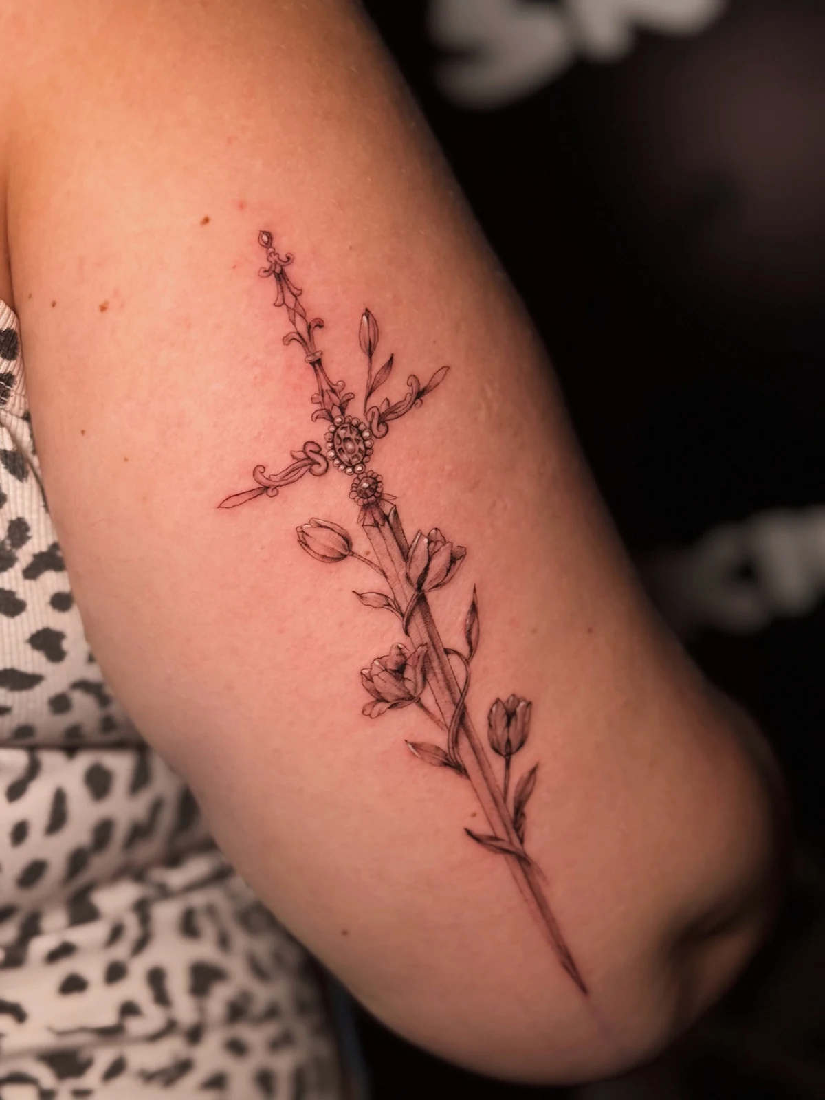 Zartes Fineline-Tattoo eines ornamentalen Kreuzes mit kleinen Blüten und Blättern am Oberarm – Arbeit von Merci, Skin City Tattoo.
