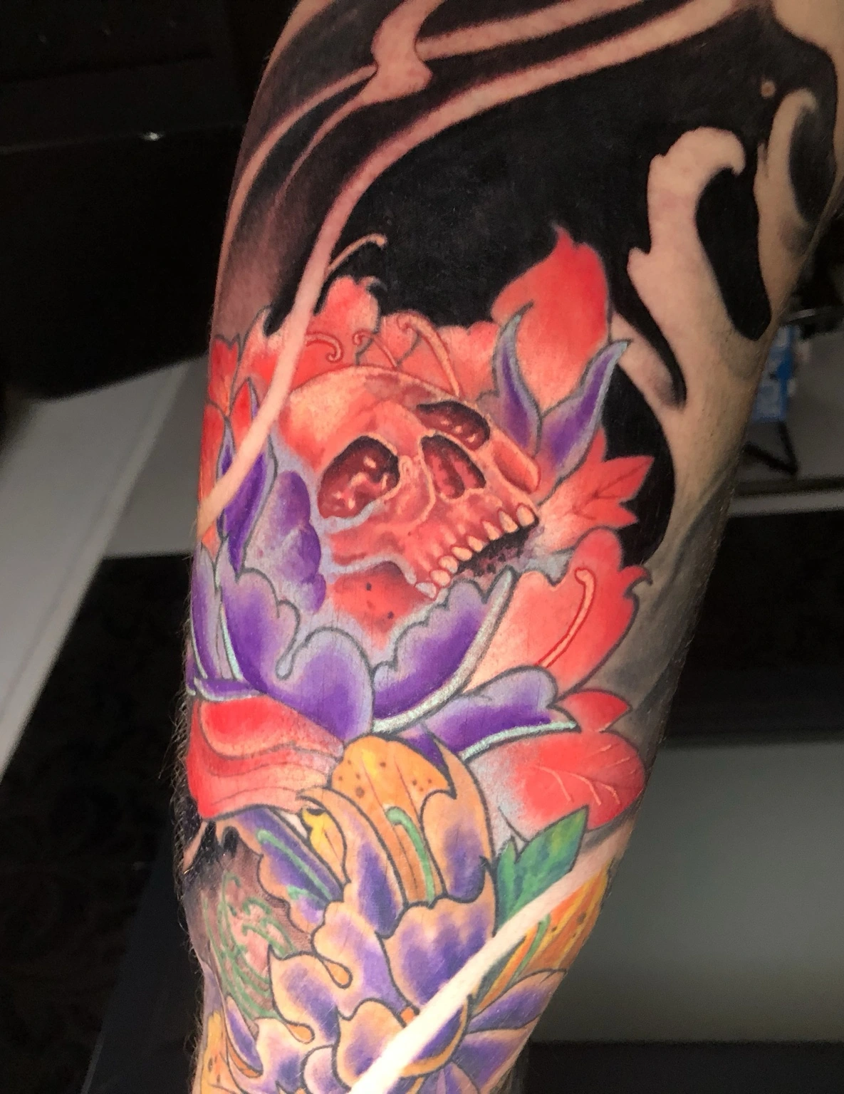 Farbiges Tattoo eines roten Schädels zwischen Blüten und schwarzen Wellen